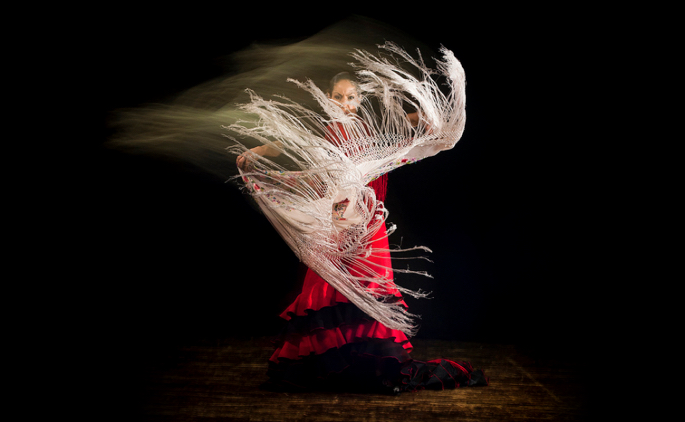 Flamenco dancer with a manton (shawl) in motion against a black background.