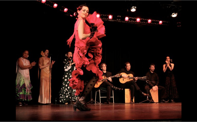 Flamenco dancer in a red dress performing footwork on stage with musicians and singer singers in the background.