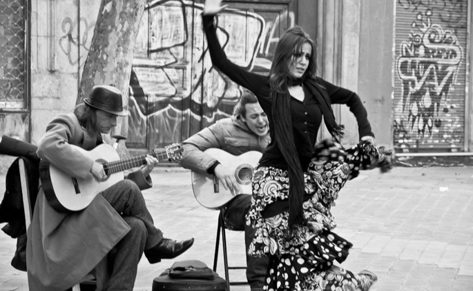 A dancer performing with two guitar players outdoors on the street with graffiti on the walls in the background.